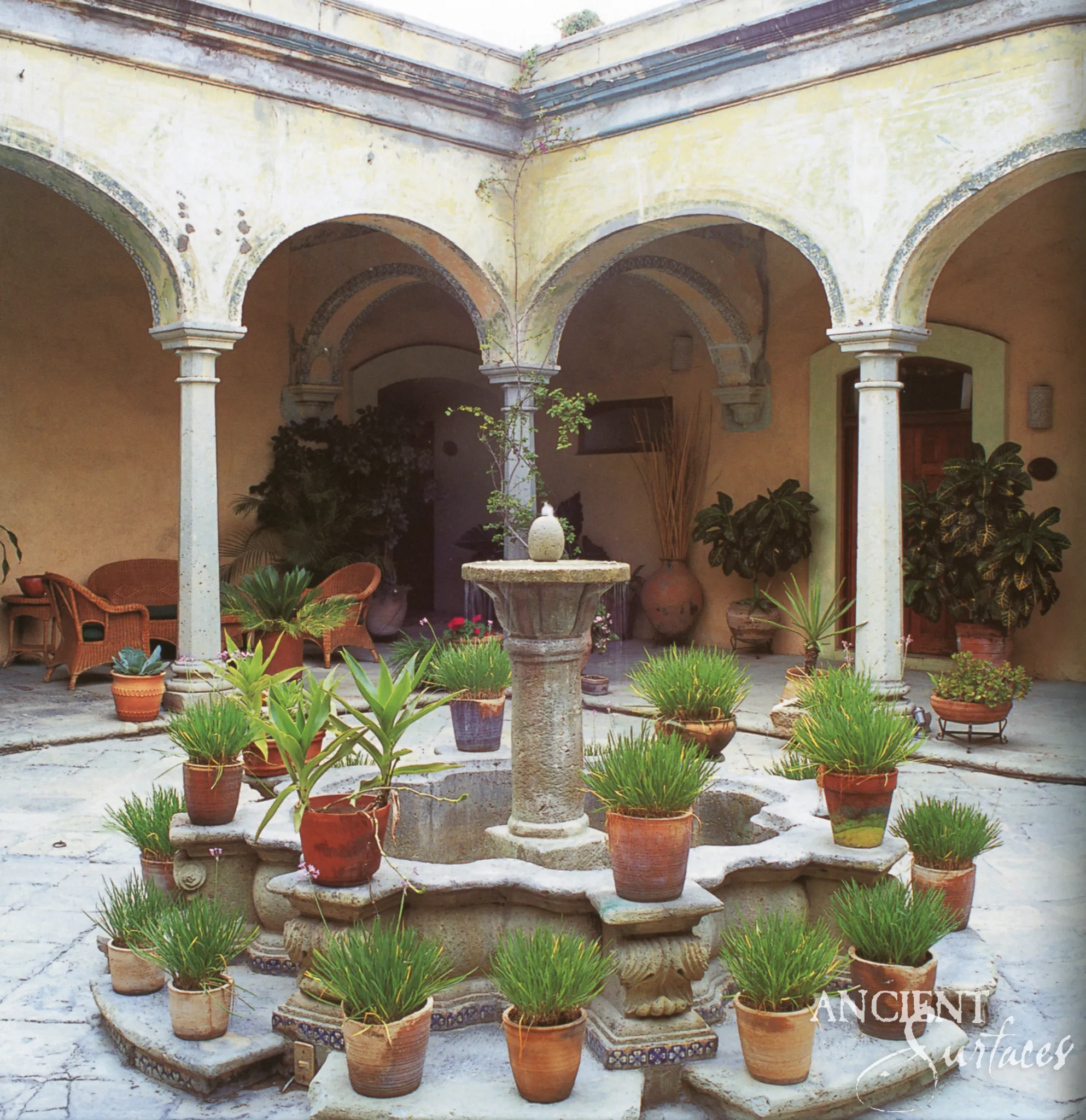 Antique limestone courtyard fountain surrounded by potted plants beneath weathered stone arches